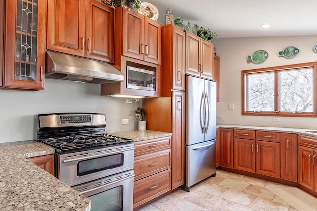 kitchen with brown cabinetry, light stone countertops, glass insert cabinets, under cabinet range hood, and appliances with stainless steel finishes