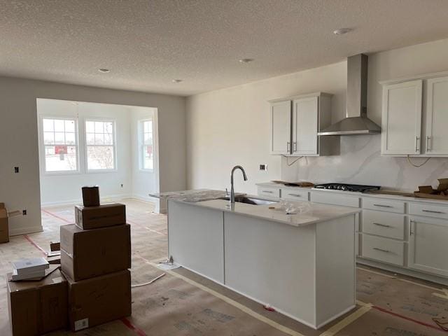 kitchen featuring wall chimney range hood, a textured ceiling, a kitchen island with sink, a sink, and stovetop