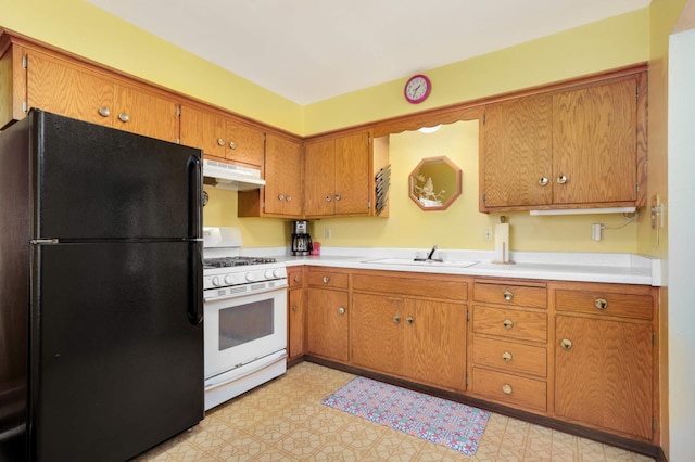 kitchen with brown cabinetry, freestanding refrigerator, white range with gas stovetop, under cabinet range hood, and a sink
