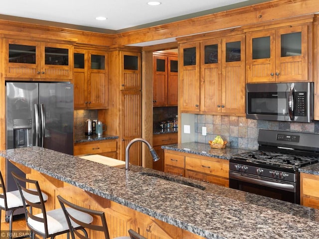 kitchen featuring dark stone counters, decorative backsplash, appliances with stainless steel finishes, a sink, and a kitchen breakfast bar
