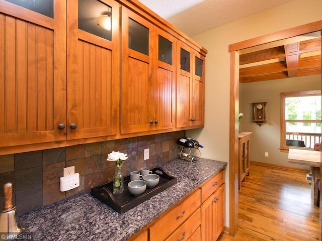 kitchen featuring dark stone counters, brown cabinets, decorative backsplash, light wood-style flooring, and glass insert cabinets