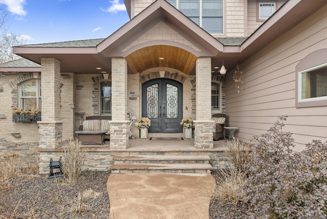 view of exterior entry with french doors, a shingled roof, covered porch, and brick siding