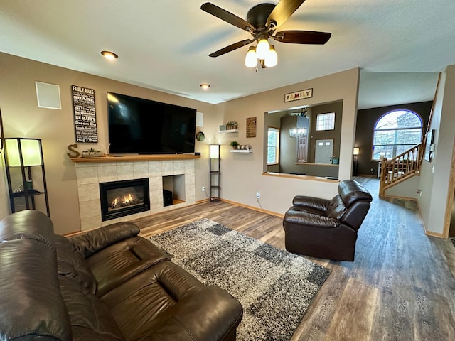 living area featuring stairs, wood finished floors, ceiling fan, a tiled fireplace, and baseboards