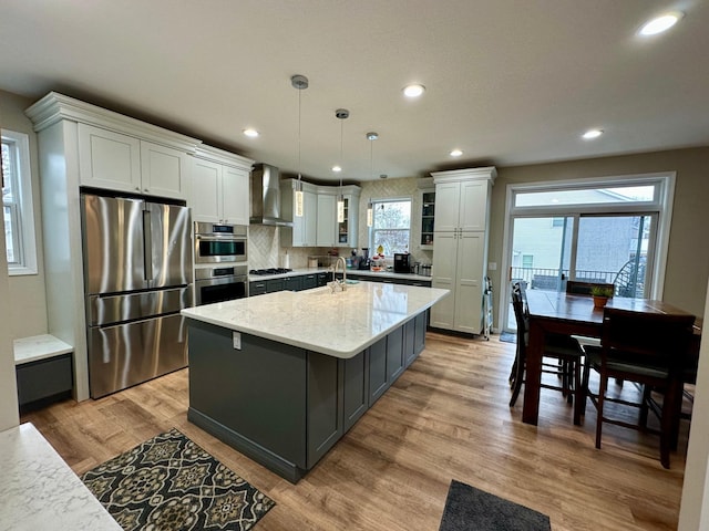 kitchen featuring stainless steel appliances, a center island with sink, recessed lighting, wall chimney range hood, and light stone countertops