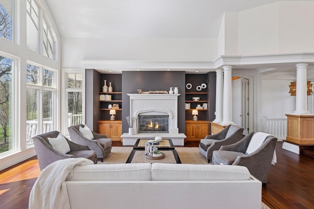 living area with a glass covered fireplace, decorative columns, built in shelves, and wood finished floors
