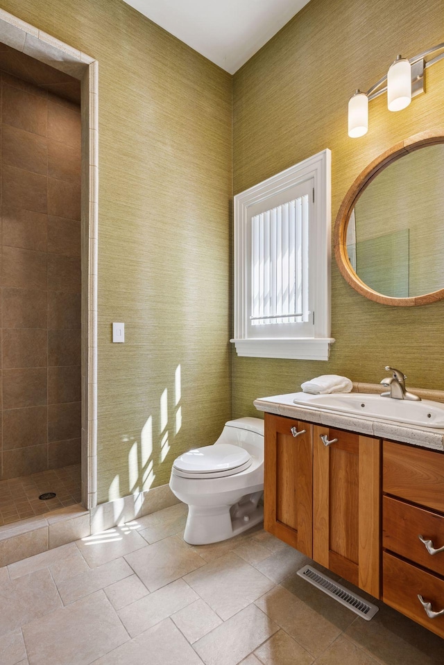bathroom with a tile shower, vanity, toilet, wallpapered walls, and visible vents