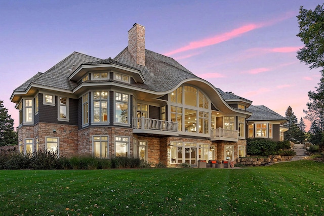 back of property at dusk with a lawn, a chimney, a balcony, and stone siding
