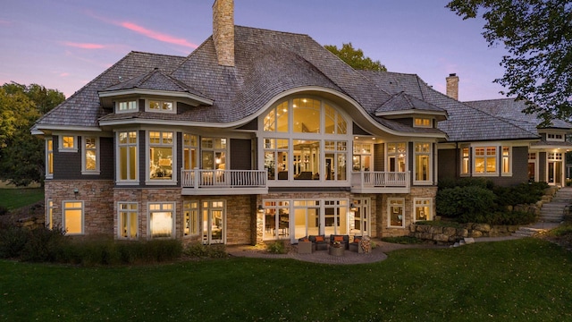 back of property at dusk with a balcony, a chimney, a yard, and stone siding