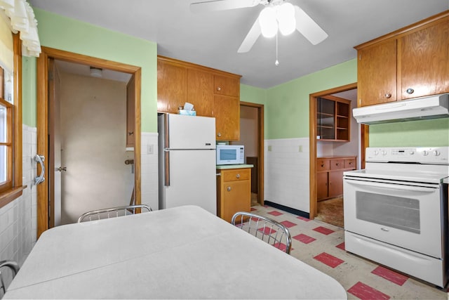 kitchen featuring brown cabinets, white appliances, ceiling fan, a wainscoted wall, and under cabinet range hood