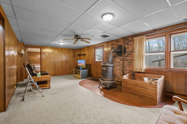 living area featuring wooden walls, ceiling fan, carpet floors, a wood stove, and visible vents
