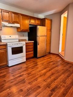 kitchen featuring under cabinet range hood, brown cabinetry, white appliances, and dark wood-type flooring