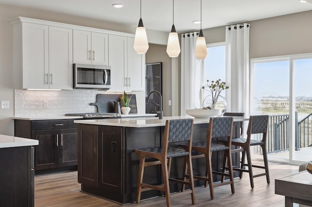 kitchen featuring stainless steel microwave, backsplash, a kitchen island with sink, light countertops, and wood finished floors