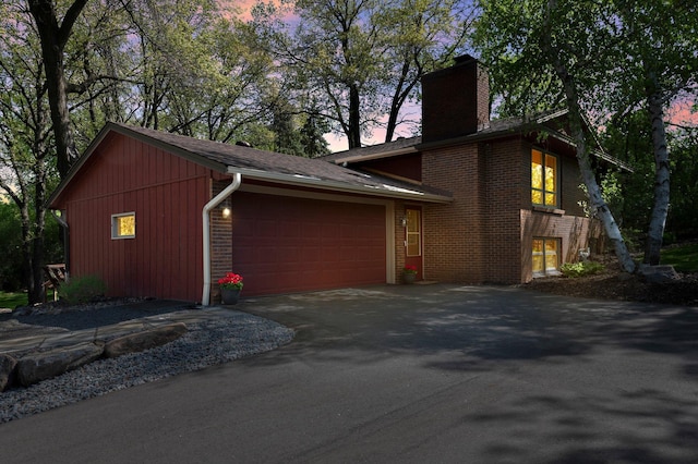 property exterior at dusk with a chimney, driveway, brick siding, and an attached garage
