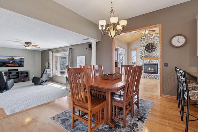 dining space with lofted ceiling, ornate columns, a stone fireplace, light wood-style floors, and ceiling fan with notable chandelier