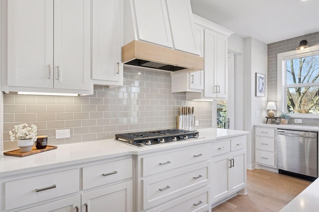 kitchen with light countertops, appliances with stainless steel finishes, light wood-style floors, custom range hood, and backsplash