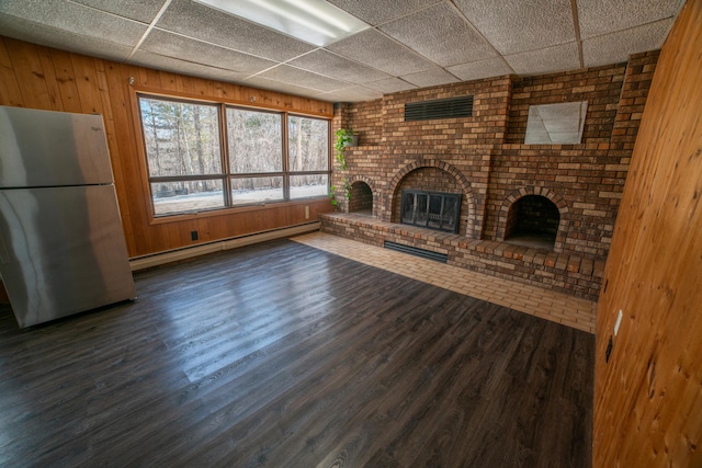 unfurnished living room with baseboard heating, a paneled ceiling, a fireplace, wooden walls, and dark wood finished floors