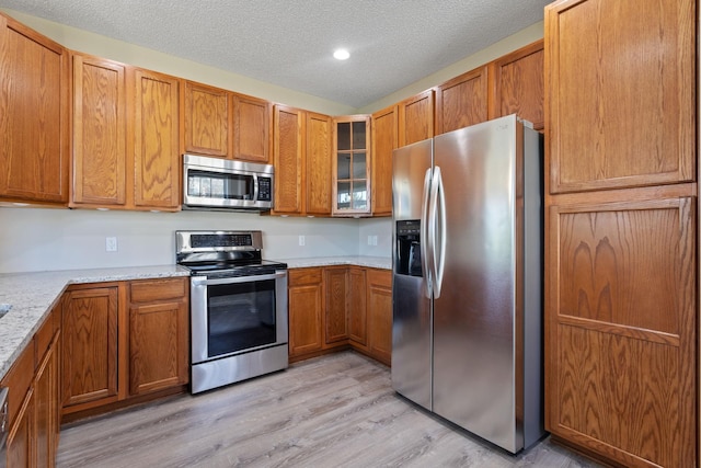 kitchen featuring appliances with stainless steel finishes, brown cabinets, light wood-type flooring, a textured ceiling, and glass insert cabinets
