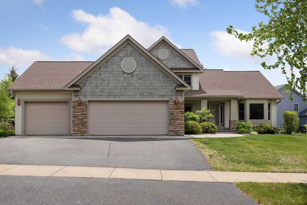 2164 Rushmore Dr, River Falls, WI 54022 - view of front facade featuring an attached garage, driveway, a front lawn, roof with shingles, and stone siding