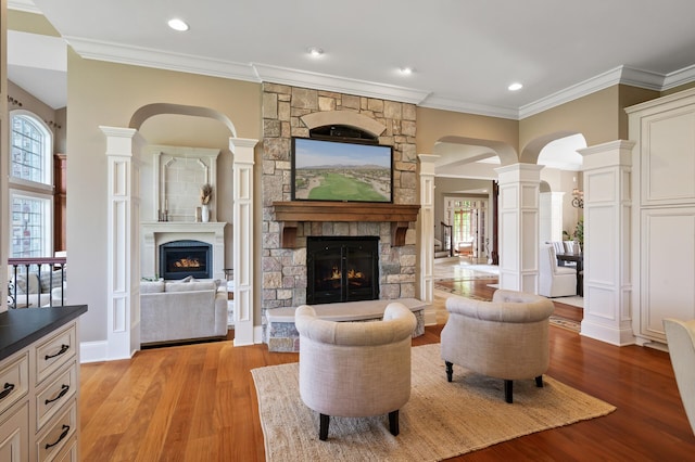 living area featuring decorative columns, arched walkways, light wood-type flooring, ornamental molding, and a stone fireplace
