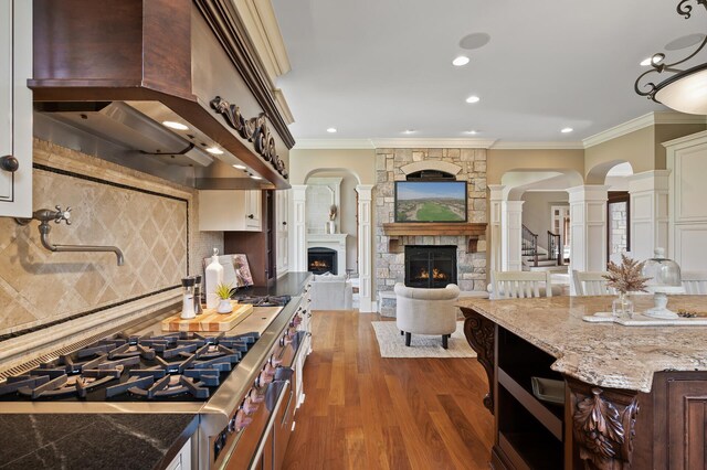 kitchen with arched walkways, ornamental molding, wall chimney range hood, a stone fireplace, and decorative columns