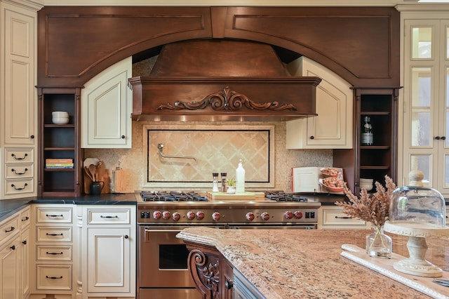 kitchen with custom exhaust hood, cream cabinetry, range with two ovens, and backsplash
