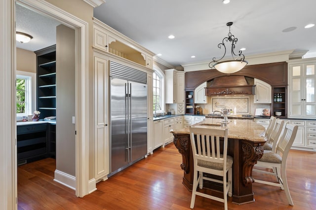kitchen featuring ornamental molding, premium range hood, built in fridge, a center island with sink, and light stone counters