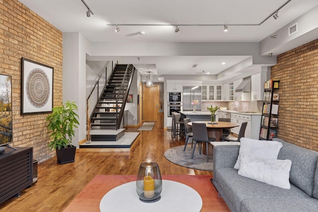 living area featuring brick wall, light wood-style floors, stairs, and rail lighting