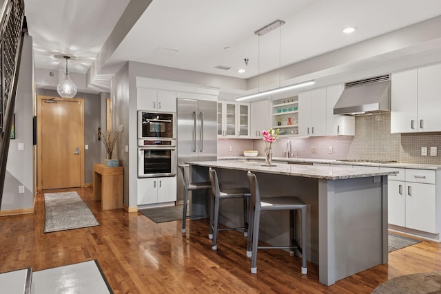 kitchen featuring ventilation hood, appliances with stainless steel finishes, backsplash, wood finished floors, and a kitchen island with sink