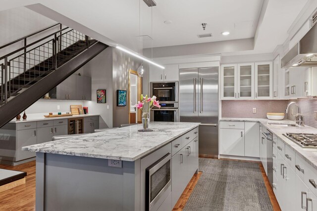 kitchen with a sink, built in appliances, wall chimney range hood, a kitchen island, and wood finished floors