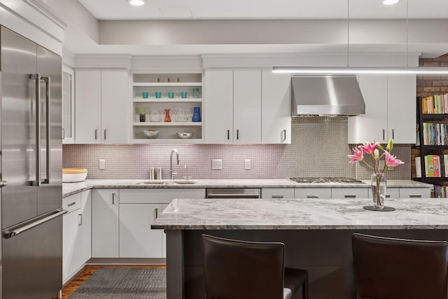 kitchen with appliances with stainless steel finishes, wall chimney exhaust hood, a sink, white cabinetry, and a breakfast bar