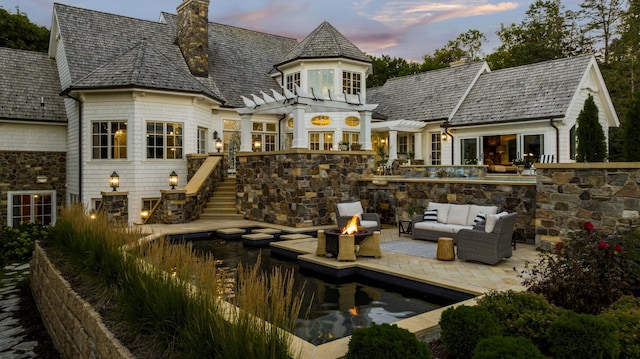 back of house at dusk with an outdoor living space with a fire pit, a patio area, stone siding, stairs, and a chimney