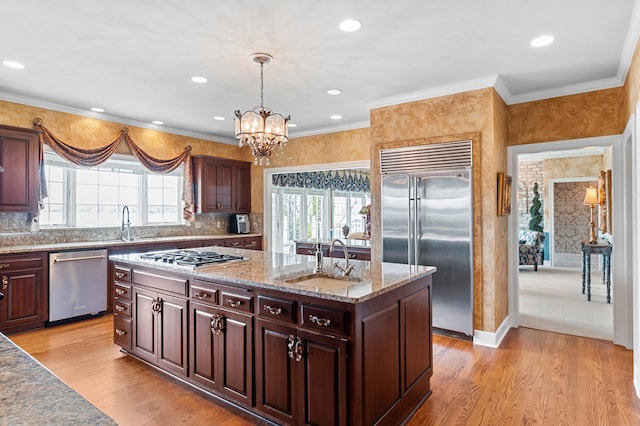 kitchen with light wood-style floors, an island with sink, a sink, appliances with stainless steel finishes, and a notable chandelier