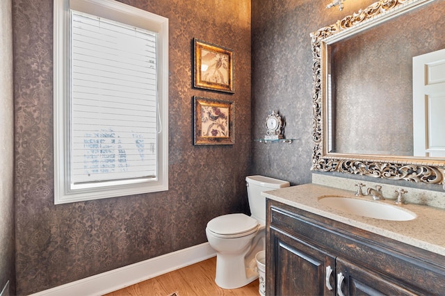 half bathroom with vanity, toilet, baseboards, wallpapered walls, and wood finished floors