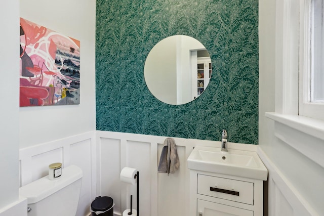 half bath featuring toilet, wallpapered walls, vanity, and wainscoting