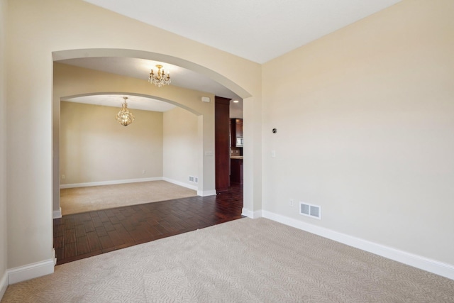 carpeted empty room with an inviting chandelier, arched walkways, visible vents, wood finished floors, and baseboards