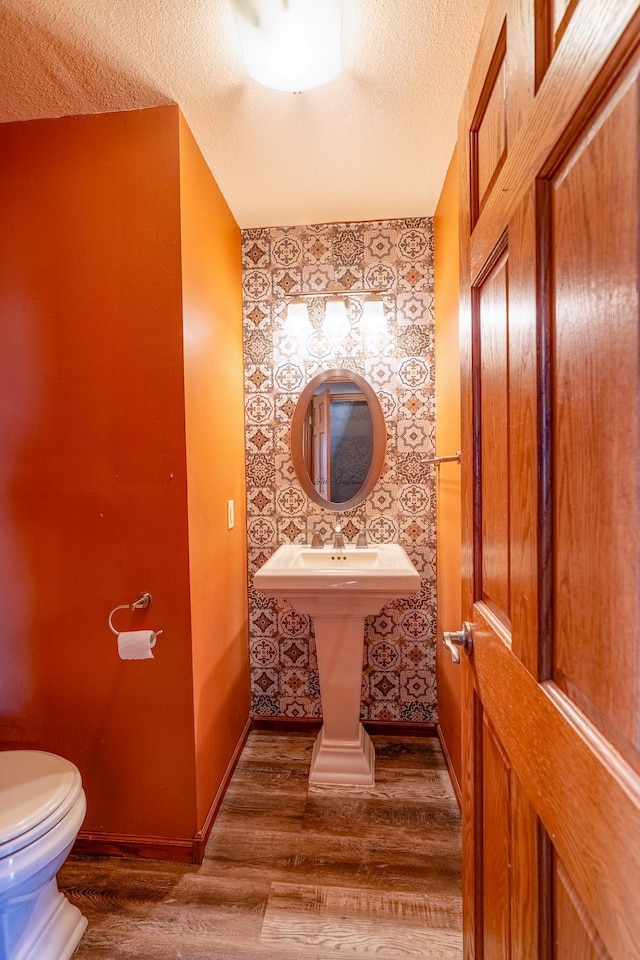 half bathroom with baseboards, wallpapered walls, a textured ceiling, toilet, and wood finished floors