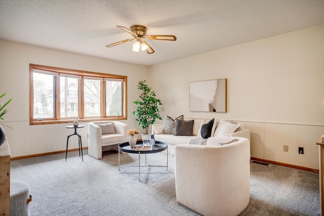 carpeted living area with a wainscoted wall, a textured ceiling, visible vents, a ceiling fan, and baseboards