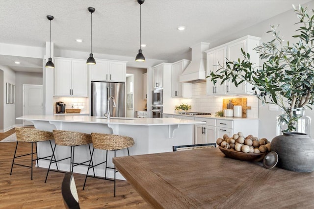 kitchen featuring appliances with stainless steel finishes, tasteful backsplash, light countertops, custom range hood, and an island with sink