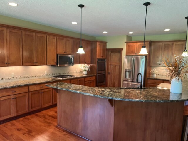 8871 Kahl Way NE, Otsego, MN 55362 - kitchen featuring appliances with stainless steel finishes, brown cabinetry, wood finished floors, a kitchen island with sink, and hanging light fixtures