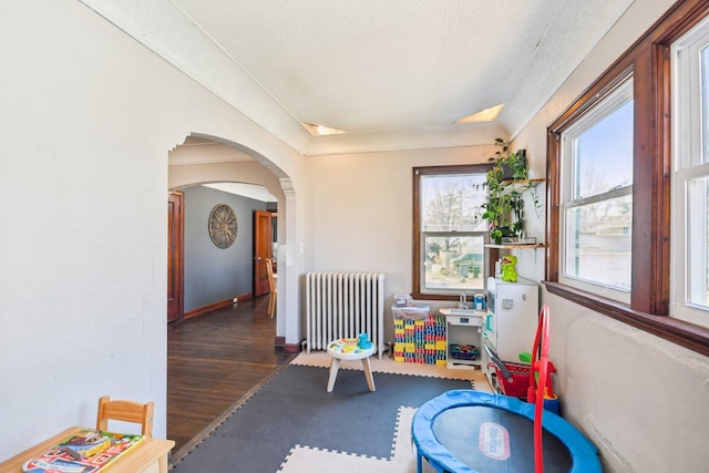 playroom with radiator, arched walkways, a textured ceiling, wood finished floors, and baseboards