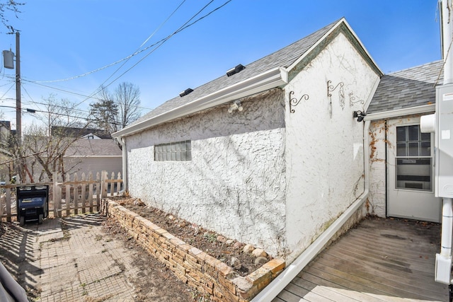 view of side of home with roof with shingles, stucco siding, a wooden deck, and fence