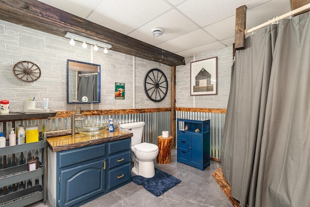 bathroom with a shower with curtain, a drop ceiling, toilet, brick wall, and vanity