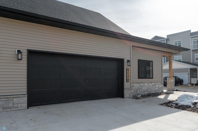 view of side of property featuring driveway, a shingled roof, stone siding, and a garage