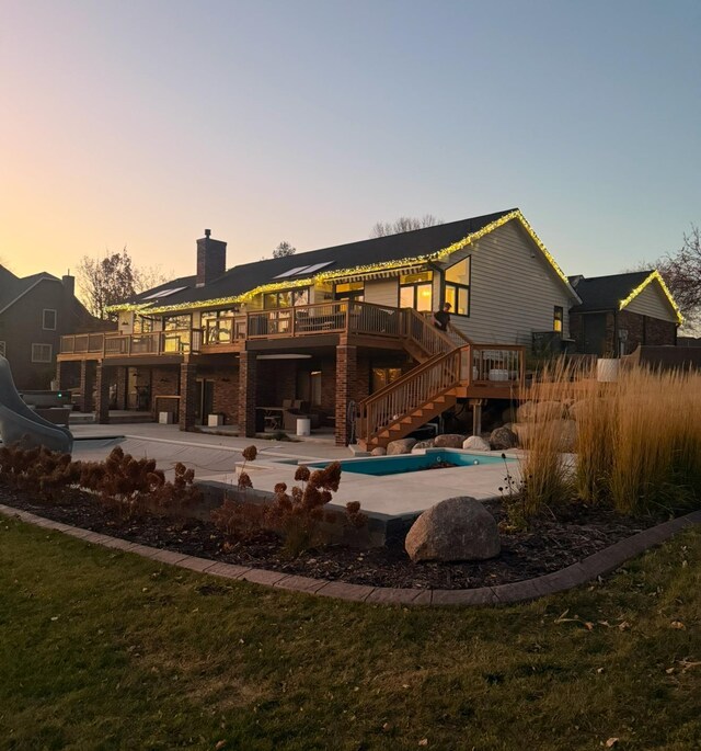 back of property at dusk with a patio area, a chimney, an outdoor pool, a wooden deck, and stairs