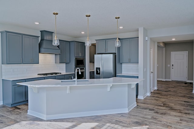 kitchen with a sink, light countertops, appliances with stainless steel finishes, premium range hood, and wood finished floors