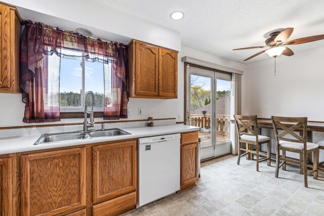 kitchen featuring white dishwasher, a sink, light countertops, brown cabinetry, and a ceiling fan