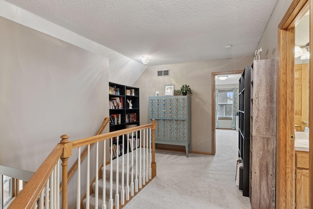 hallway featuring an upstairs landing, visible vents, baseboards, a textured ceiling, and light colored carpet