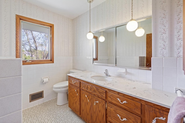bathroom featuring wallpapered walls, toilet, vanity, wainscoting, and tile walls