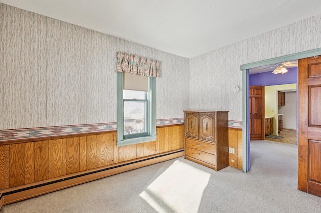 carpeted empty room featuring a baseboard heating unit, wallpapered walls, wooden walls, a ceiling fan, and a wainscoted wall