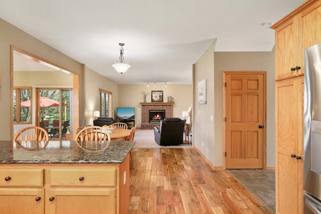 kitchen featuring dark stone counters, decorative light fixtures, stainless steel fridge, light wood finished floors, and a brick fireplace